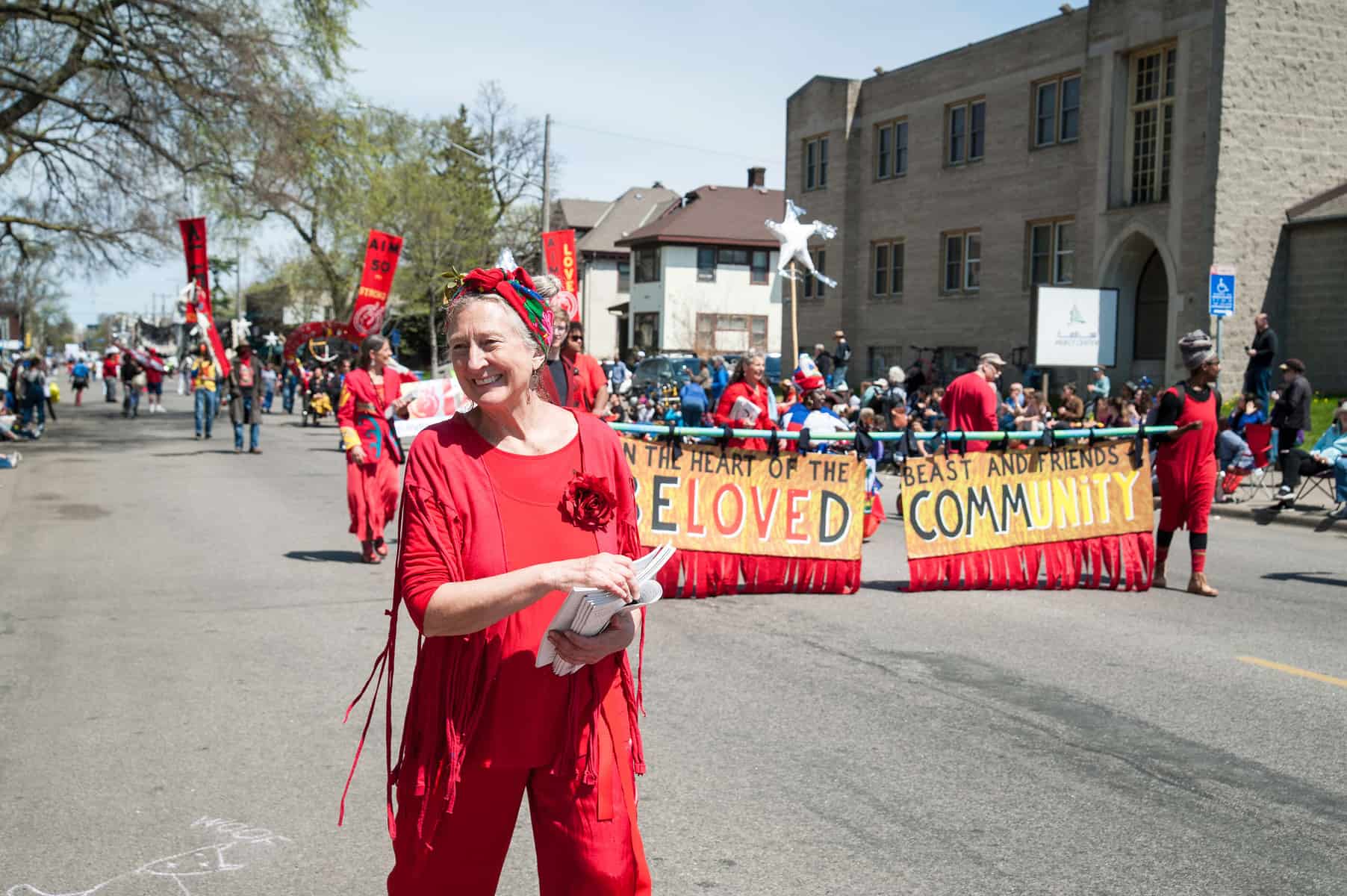 Image Description: A smiling older white person wearing all red stand in front of a group of people holding a banner. 