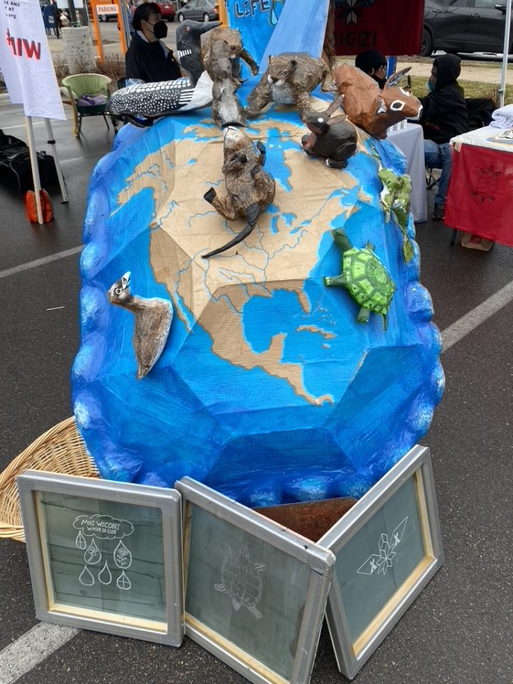 Image Description: A photo of MIGIZI's turtle puppet painted as the earth with small animal sculptures attached and three screen prints in front.