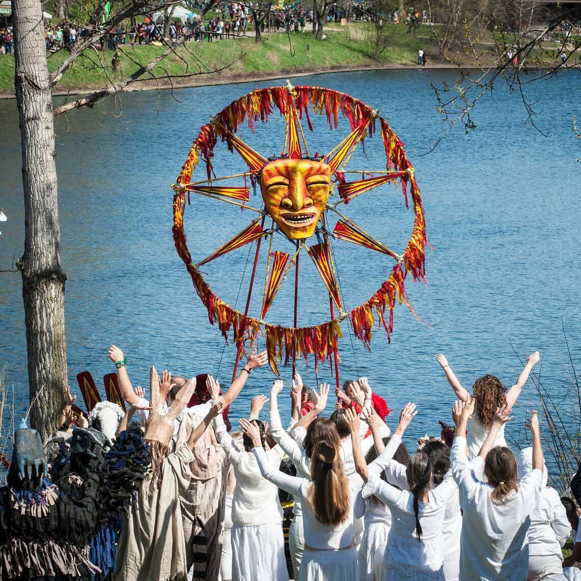 Image Description: A group of people stand together raising their arms. A few of them are holding up a large puppet that is a sun with a face. They are all in front of a lake.