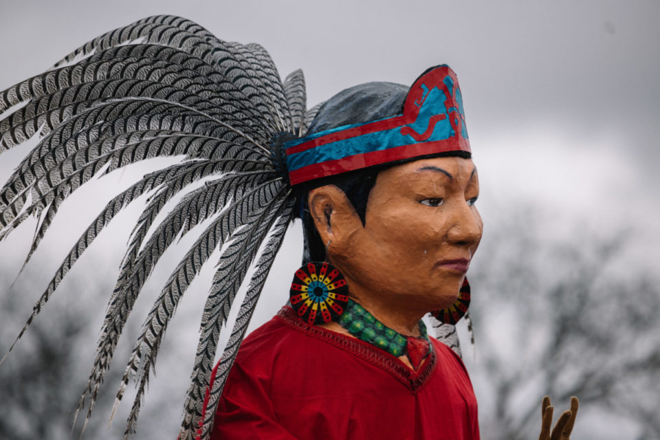 Image Description: A photo of Kalpulli KetzalCoatlicue's Medicine Woman Mojiganga puppet standing with striped feathers tucked in a blue and red headband, large circle earrings and a green necklace, wearing red.