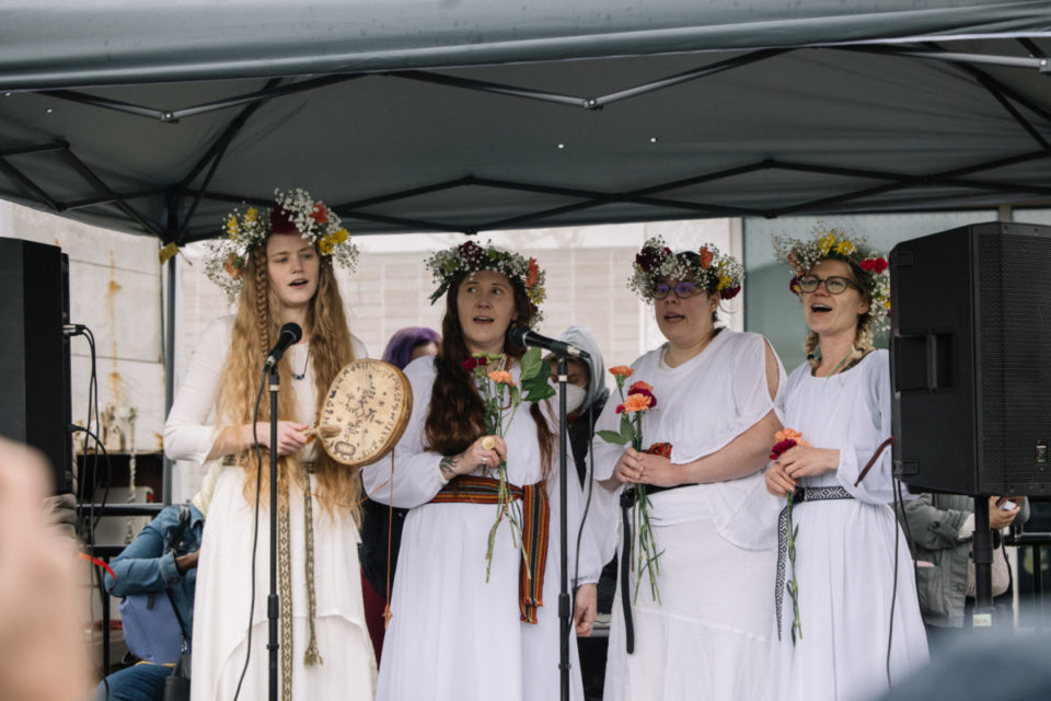Image Description: A photo of the Daughters and Sons of Yta, Scandinavian dancers sing on stage dressed in white with flower crowns holding flowers and instruments.