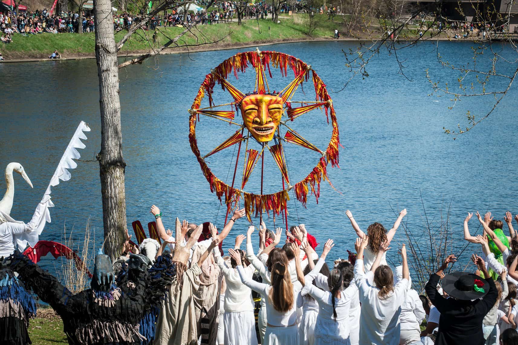 Image Description: A group of people stand together raising their arms. A few of them are holding up a large puppet that is a sun with a face. They are all in front of a lake.